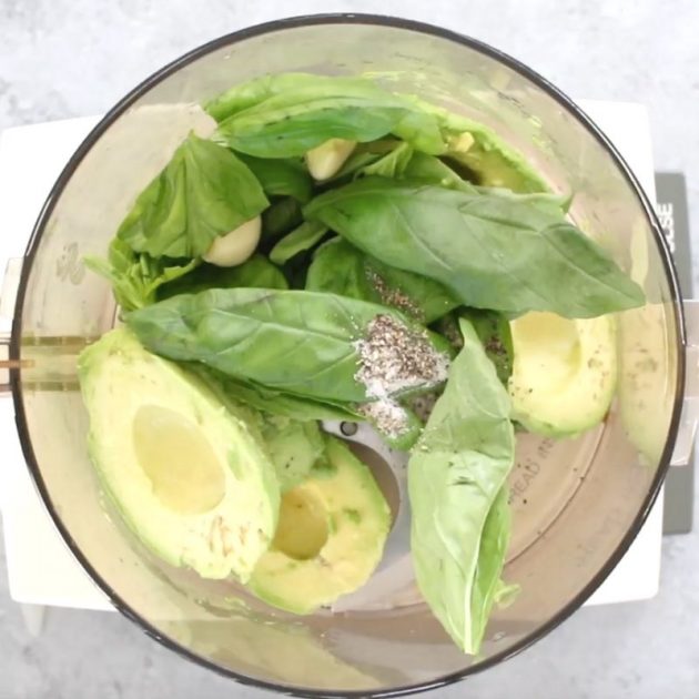 This photos shows the ingredients for avocado pasta inside a food processor: avocados, fresh basil, garlic, salt and pepper
