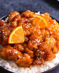 A serving of Chinese Orange Chicken garnished with sesame seeds and orange wedges on a bed of steamed rice
