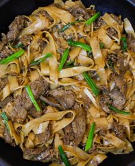 Beef chow fun noodles in a wok after being prepared showing tender strips of beef and fresh green onions for garnish