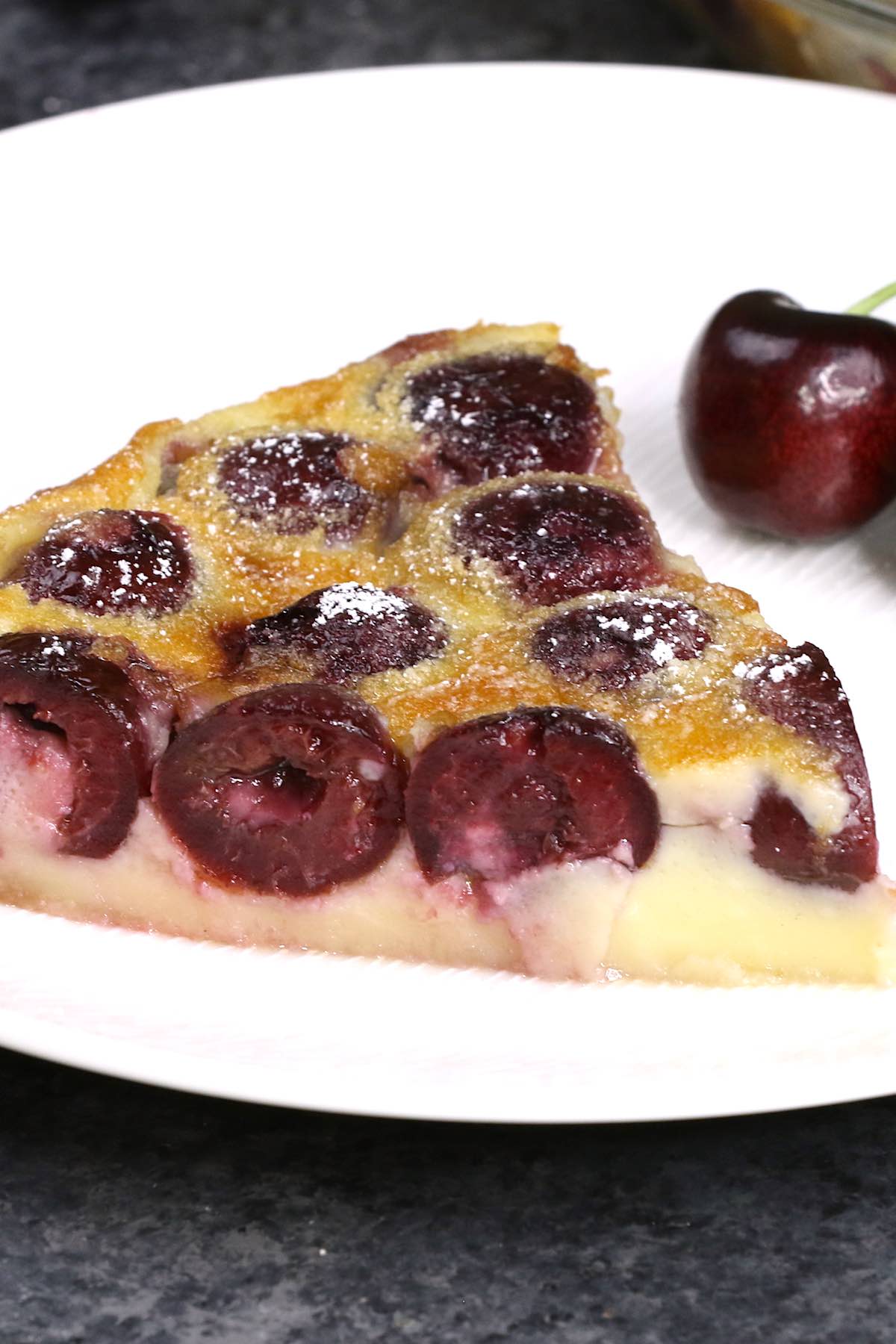 A wedge of clafoutis dusted with powdered sugar on a serving plate with a fresh cherry on the side