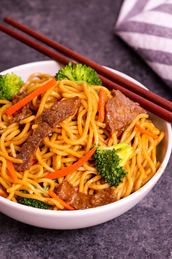 A serving of beef lo mein in a white serving bowl with chopsticks ready to be eaten