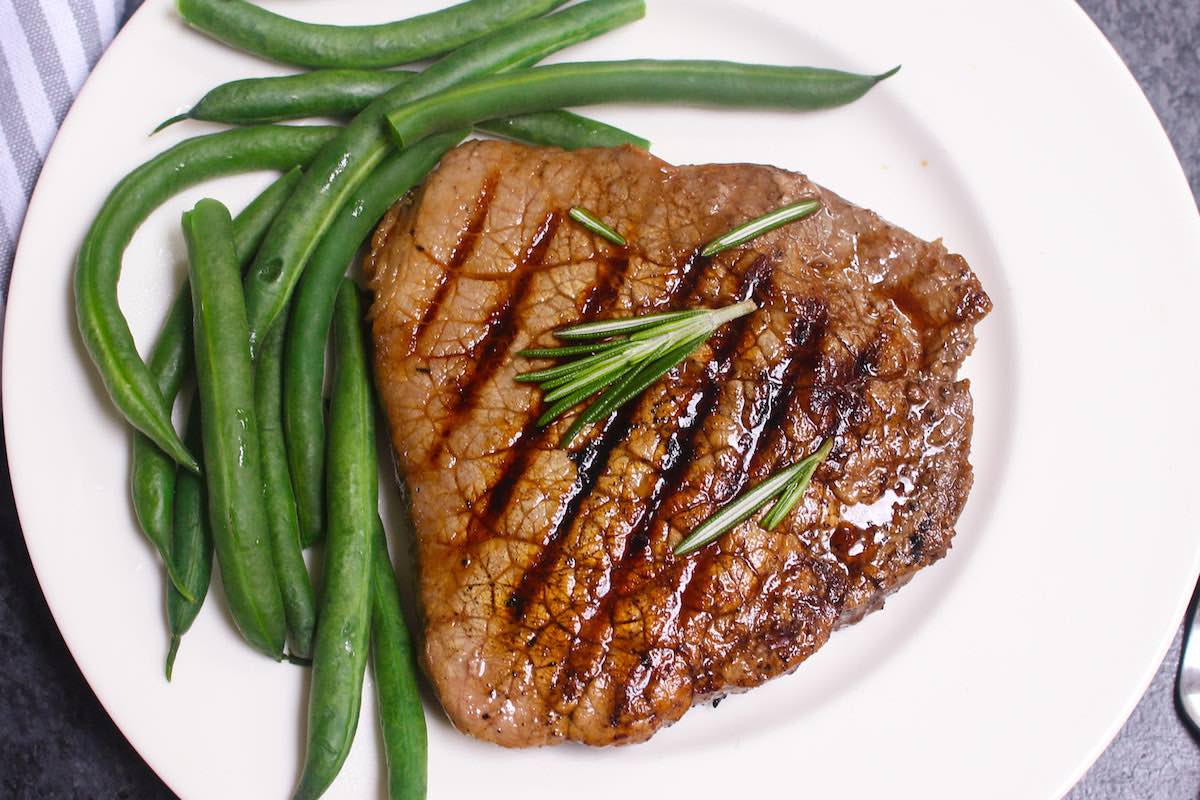 Overhead view of a grilled round steak on a serving plate with green beans as an example of a tougher cut made tender using marinating