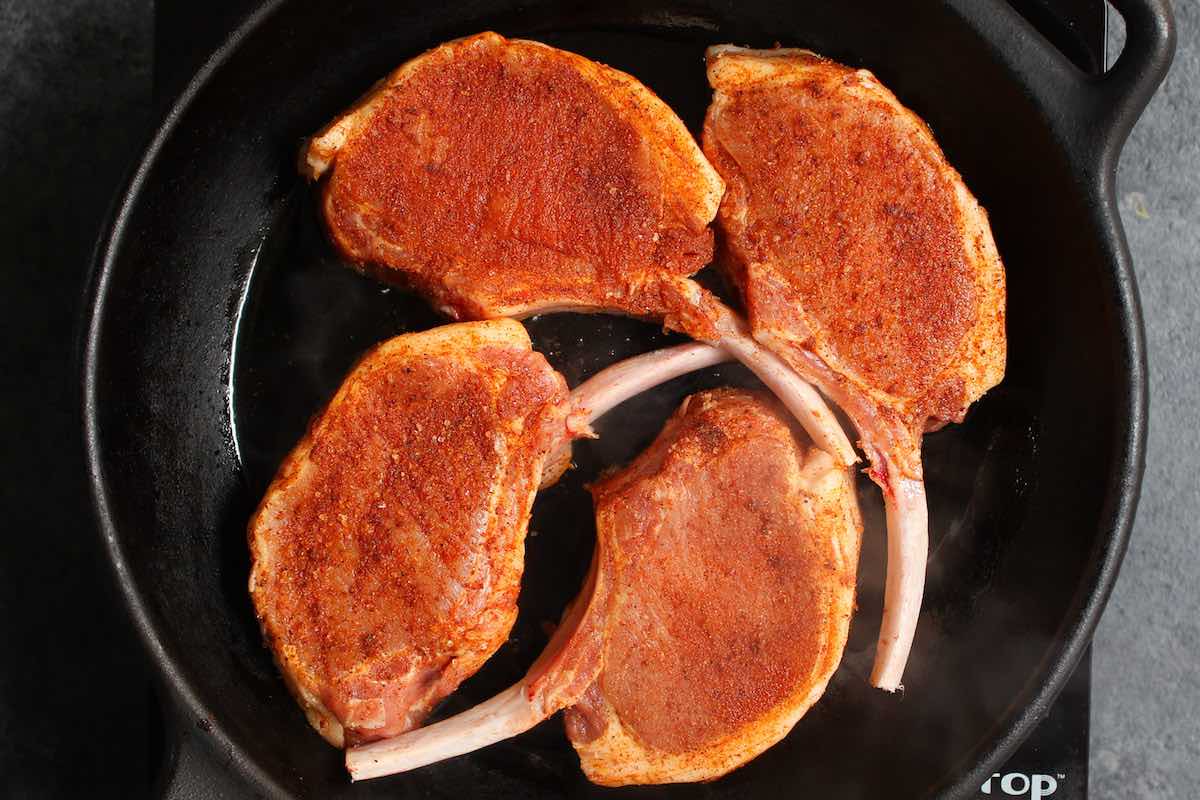 Overhead photo showing four seasoned bone-in rib chops frying in a cast iron skillet