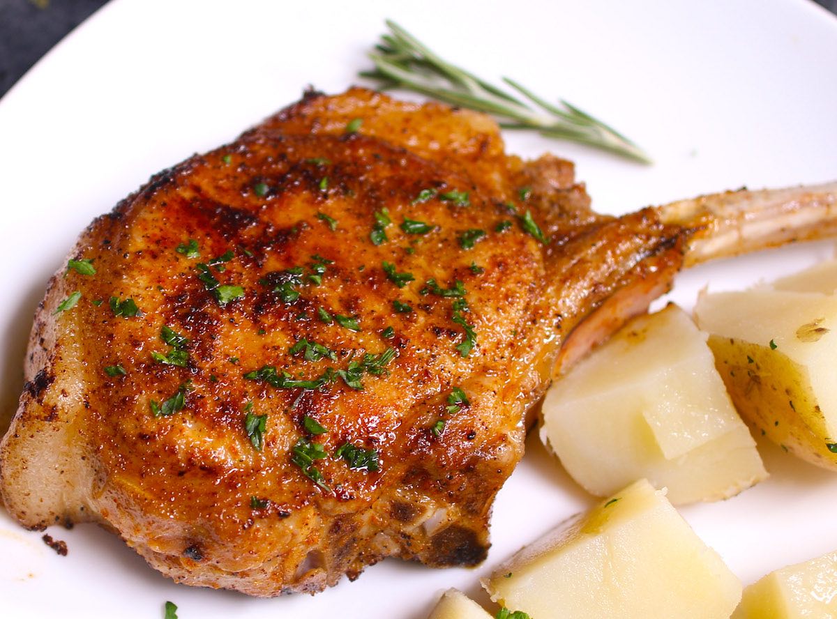 A pan fried pork chop cooked to perfection and garnished with fresh parsley and potatoes on the side on a serving plate