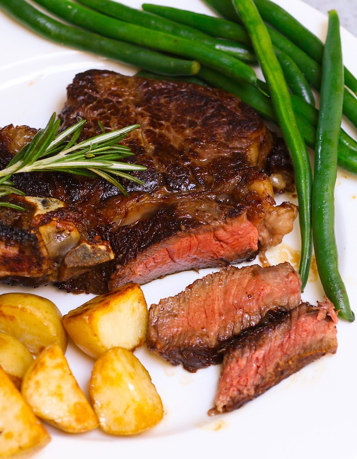 Closeup of sliced rib eye steak that has been perfectly cooked to medium rare doneness and served with green beans, sauteed potatoes and a sprig of fresh rosemaryon the side