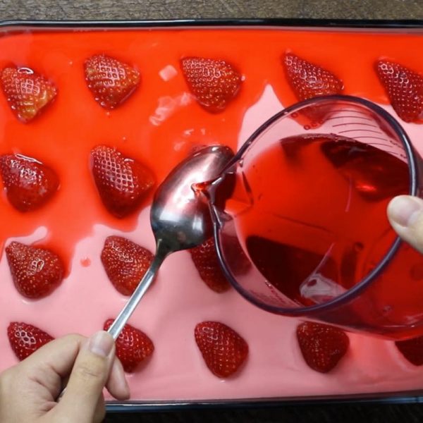 This is a photo showing how to pour the strawberry jello glaze onto the back of a spoon so it dribbles gently onto the filling layer in strawberry jello cake