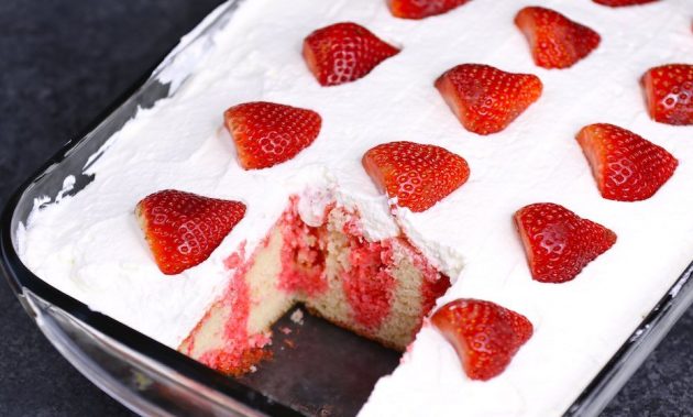 Strawberry Jello Poke Cake in the Pan after the first piece has been served, showing the arrangement of fresh strawberries on top along with the smooth cream layer and strawberry jello permeating into the poke holes in the cake